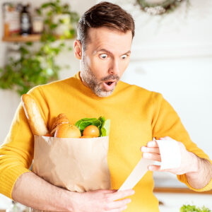Man carrying a bag of groceries and looking at bill, shocked by the price